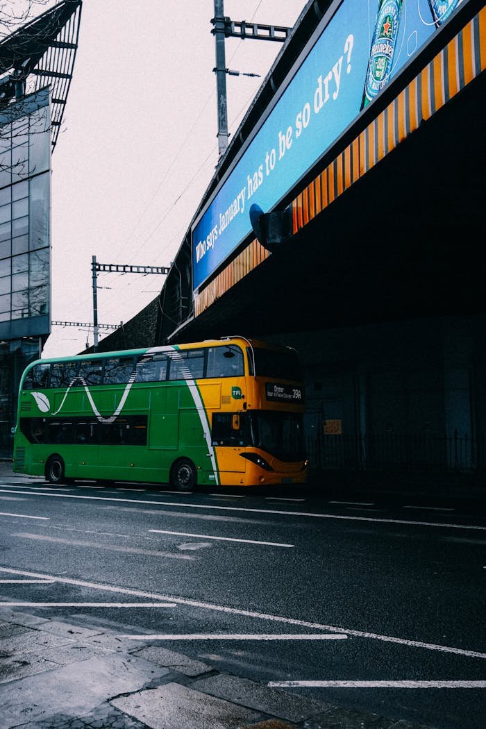 A green double-decker bus travels under a bridge in an urban setting with a poster above.