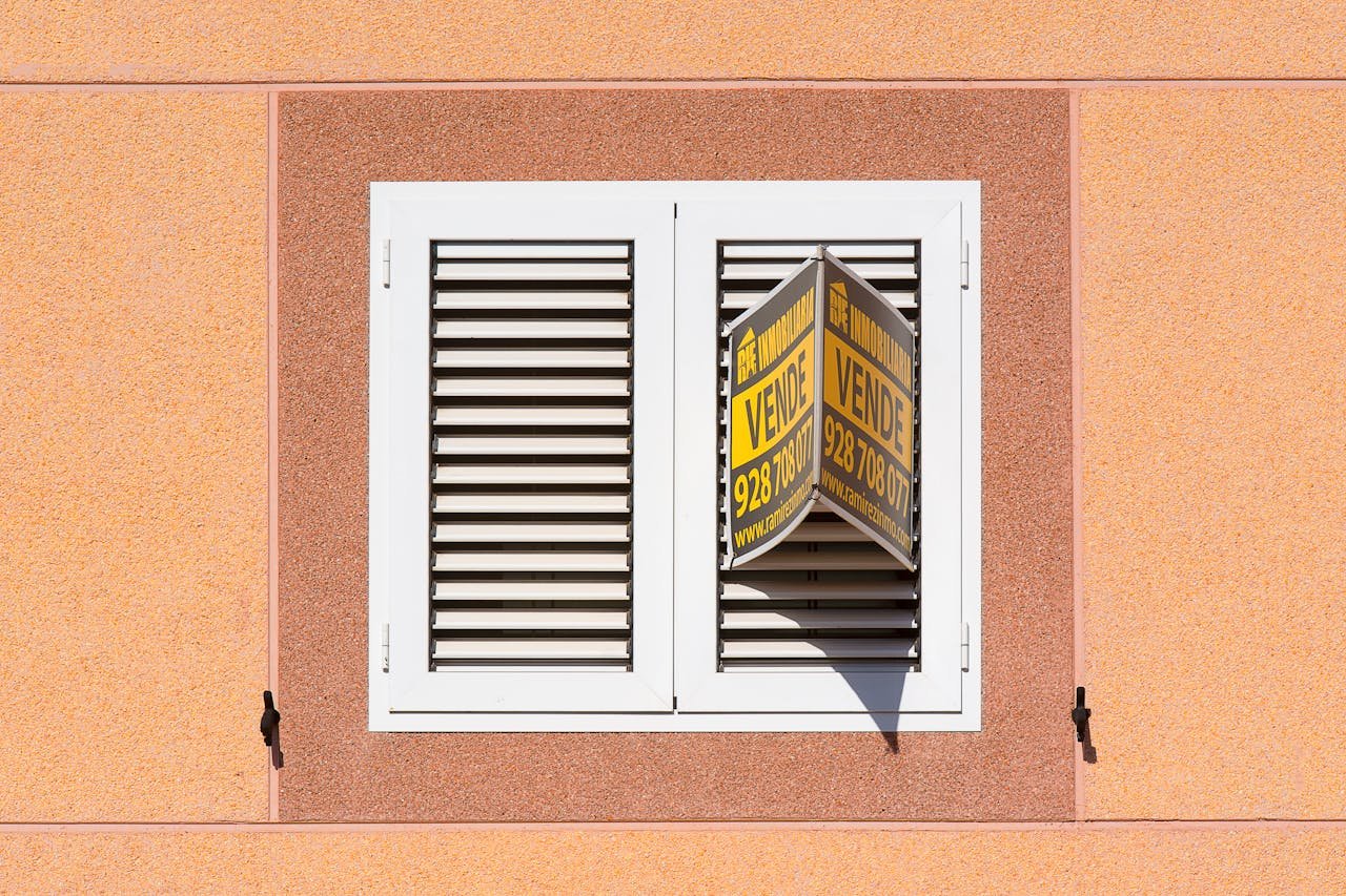 White shutter window with a for sale sign on peach building exterior.